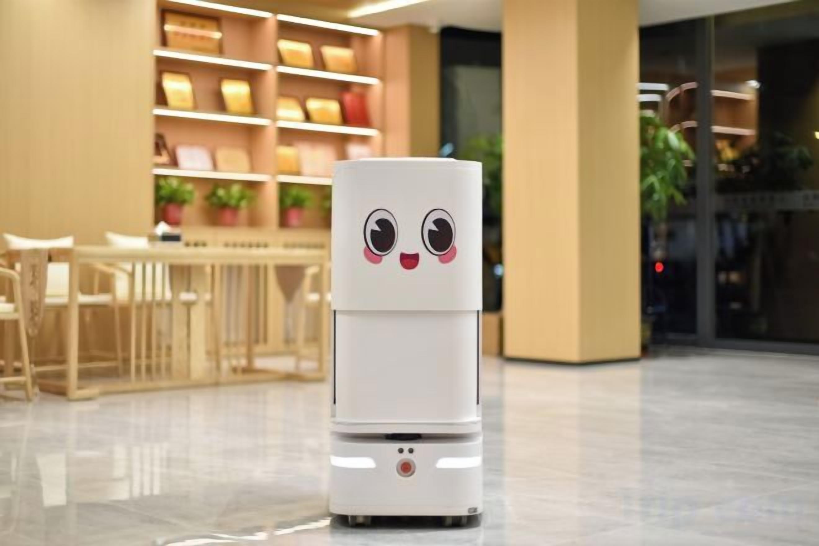 A white robot with a face in a modern indoor setting with a dining area and shelves in the background.