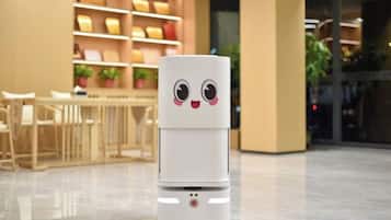 A white robot with a face in a modern indoor setting with a dining area and shelves in the background.