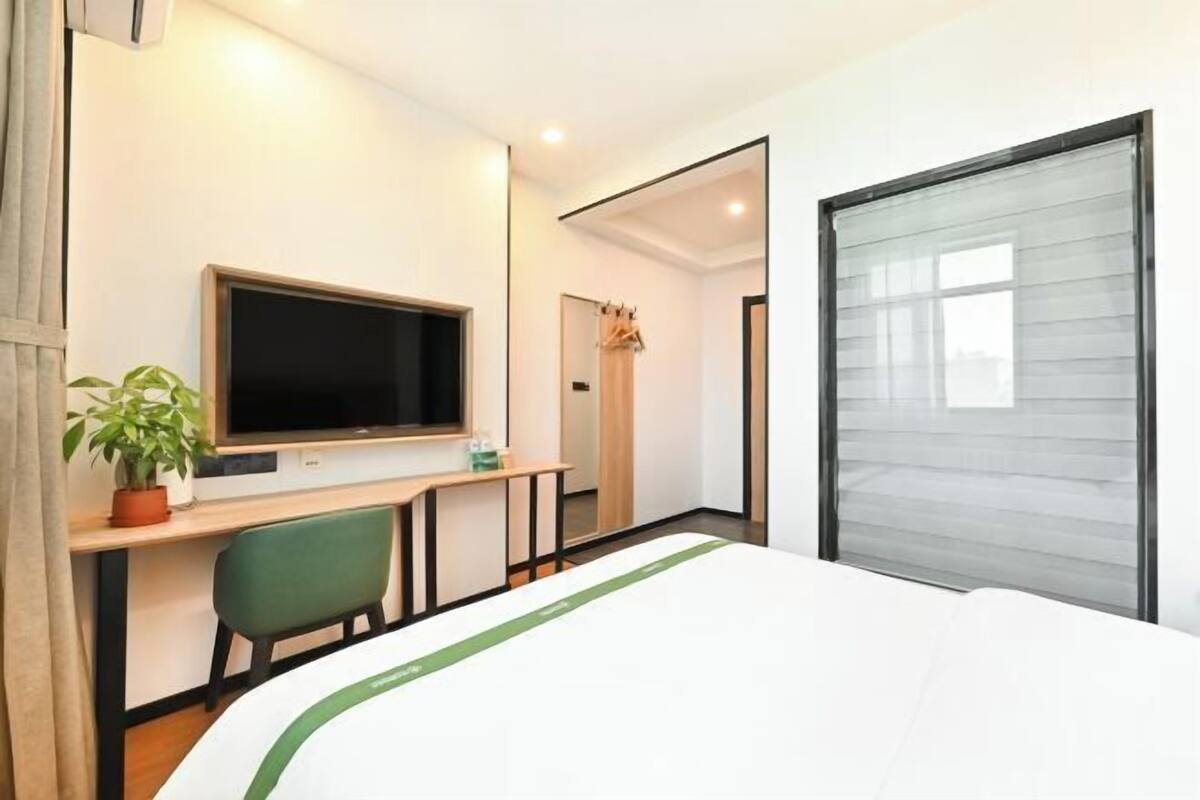 A hotel room with a bed, a desk with a TV, a chair, a plant, and a window.