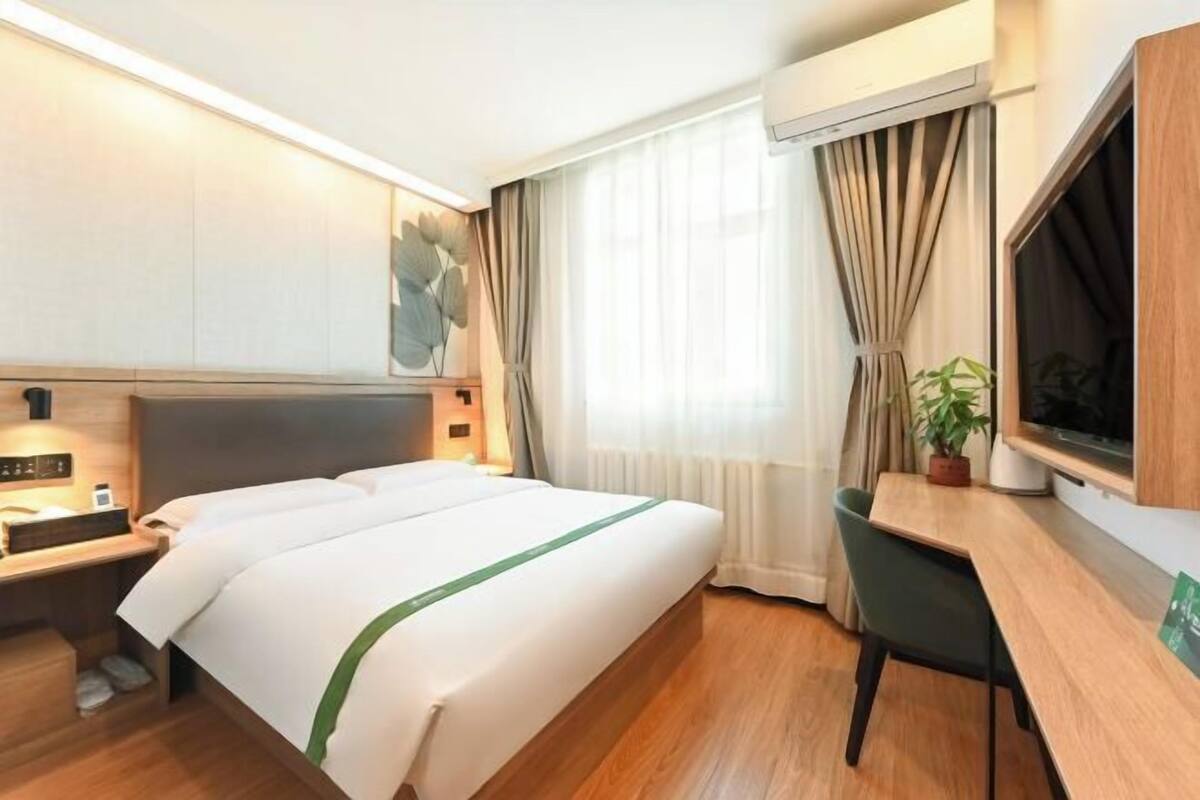 A hotel room with a bed, bedside table, TV, desk, chair, and a large window with curtains.