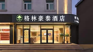 Entrada de hotel com placa em caracteres chineses, porta de vidro e interior visível com prateleiras e plantas.
