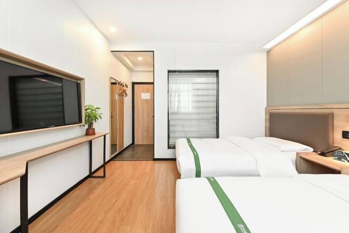 A modern hotel room with a large flat-screen TV, a wooden floor, a bed with white bedding and green stripes, a bedside table, and a window with blinds.