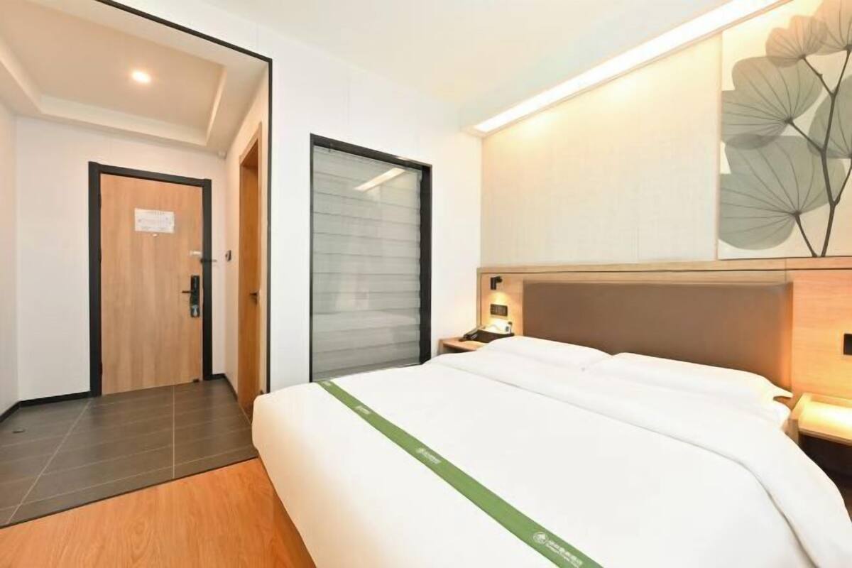 A modern hotel room with a large bed, a wooden door, a sliding glass door, and a wall-mounted artwork.