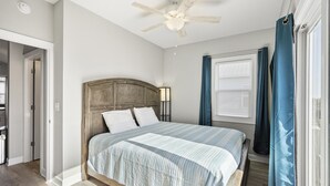 Une chambre avec une tête de lit en bois, un lit avec une literie à rayures, un ventilateur de plafond, une fenêtre avec des stores et une porte donnant sur une autre pièce.
