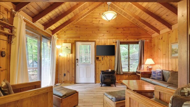 A cozy cabin interior with wooden walls and floors, a fireplace, a television, and a seating area with a sofa and benches. - Cozy Lake Cabin #3 w/boatdock (Palmyra Township)