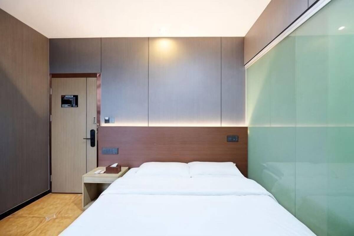 Une chambre d’hôtel comprenant un lit, une table de chevet, une commode, une tête de lit en bois et une cloison en verre.