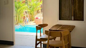 Een kamer met een houten tafel en twee stoelen, een houten deur en uitzicht op een zwembad en palmbomen door een boogvormige deuropening.