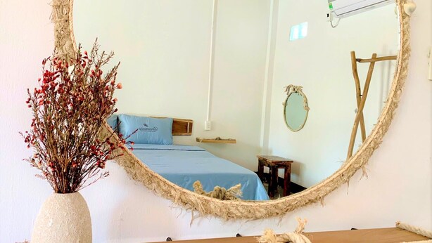 Een slaapkamer met een grote spiegel, een bed met blauw beddengoed, een klein houten krukje en een rustieke touwdecoratie.
