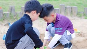 Dos niños jugando en una zona arenosa, con postes de madera al fondo.