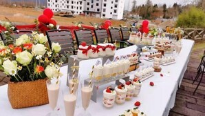 Una mesa de buffet con diversos postres y bebidas, ubicada al aire libre con sillas y un edificio al fondo.