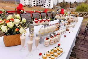 Una mesa de buffet con diversos postres y bebidas, ubicada al aire libre con sillas y un edificio al fondo.