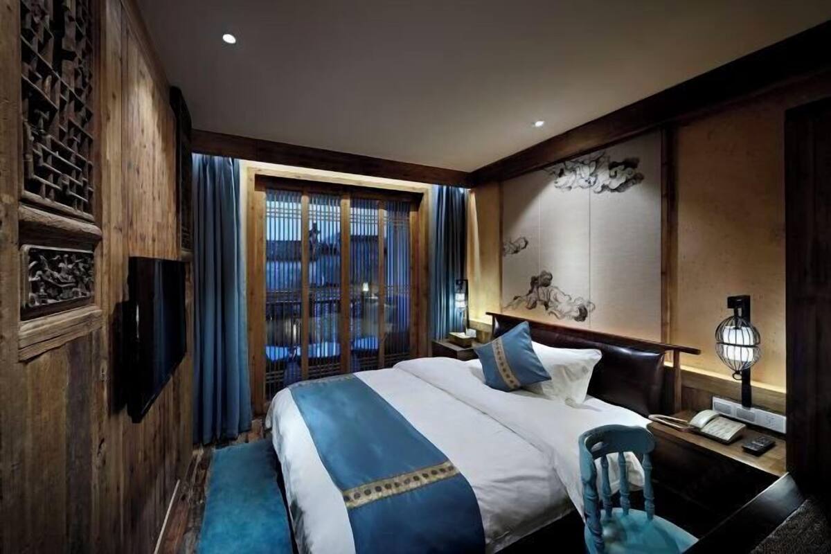 A hotel room with a large bed, a desk with a chair, a TV mounted on the wall, and a window with curtains.