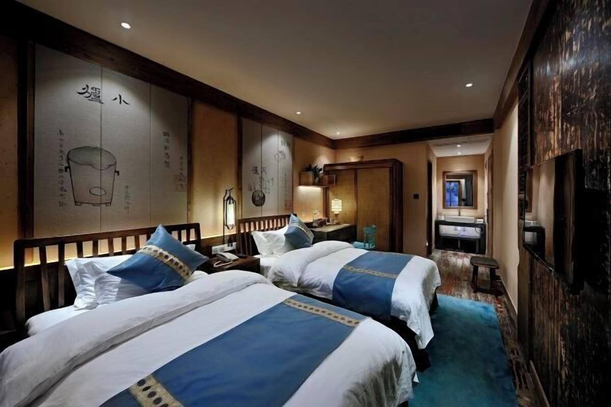 A hotel room with two beds, a wooden headboard, a wall with calligraphy, a small table with a lamp, and a bathroom visible through an open door.