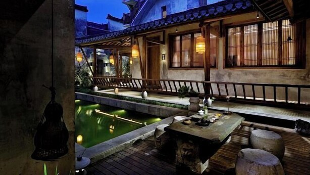 A traditional courtyard with a wooden table and benches, a small pond, and a building with tiled roofs and wooden railings.