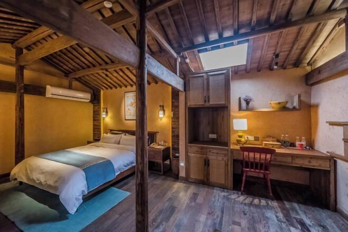 A rustic room with a bed, desk, and chair, wooden beams on the ceiling, and a window.