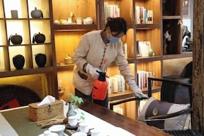 Una persona limpiando una mesa de madera con un pulverizador en una habitación con estanterías llenas de libros y cerámica.
