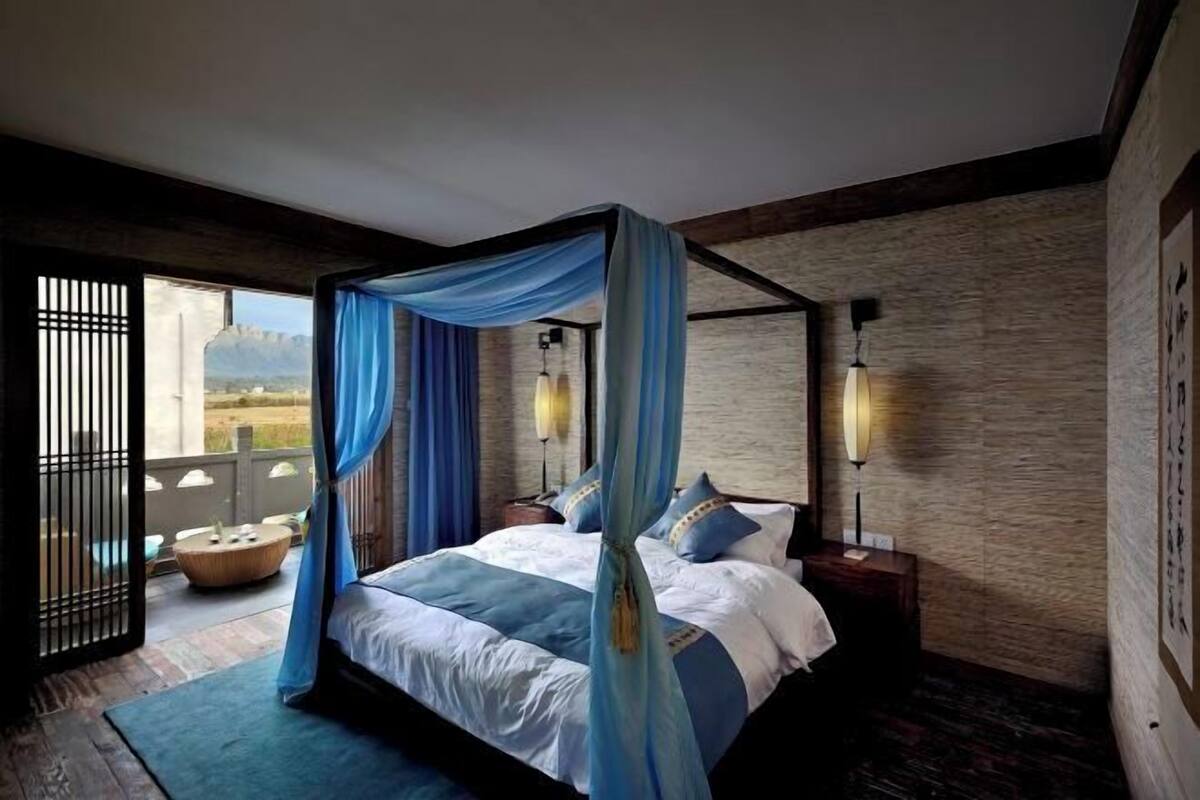 A bedroom with a four-poster bed, a view through a sliding door, and a nightstand with a lamp.
