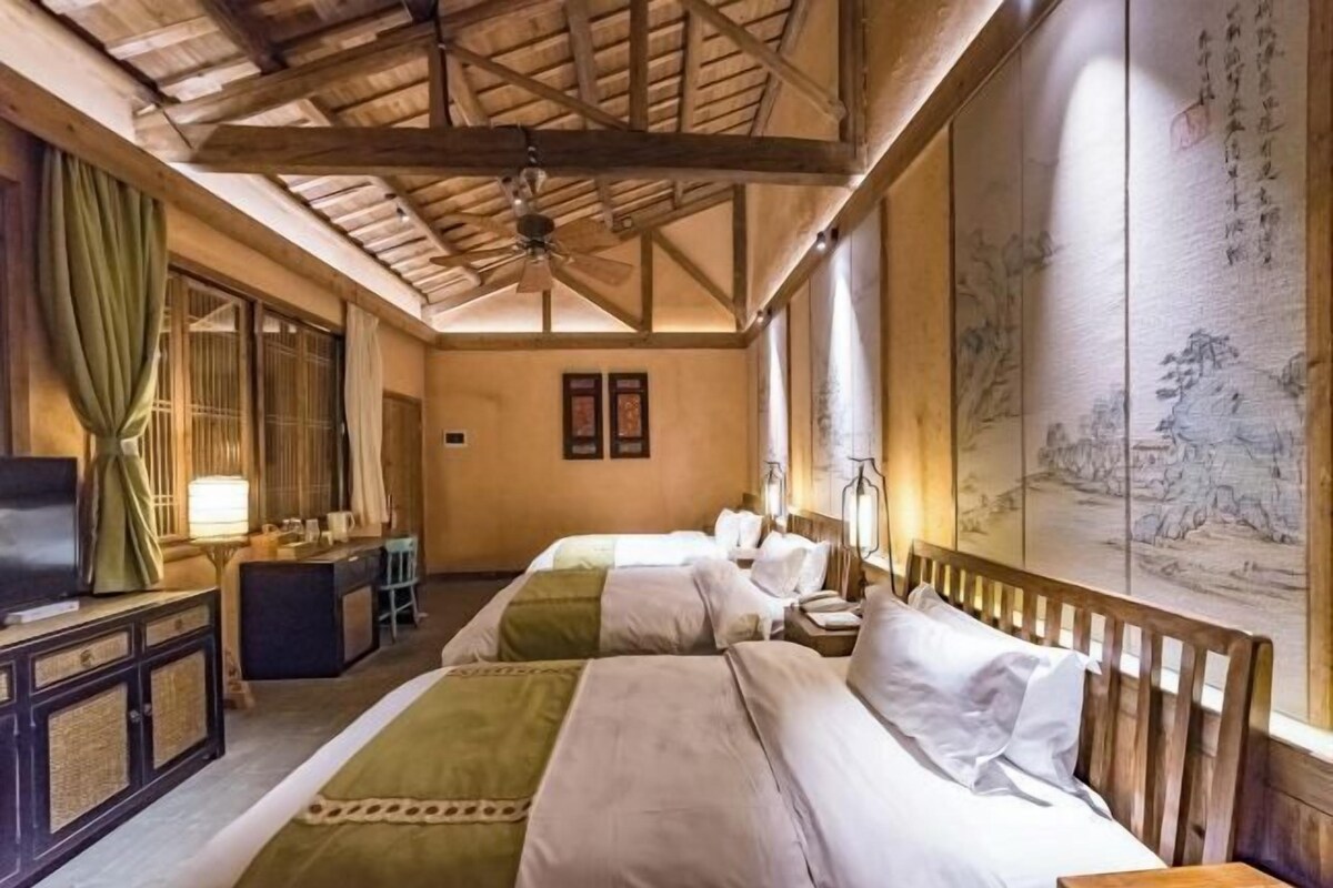 Een hotelkamer met een houten plafond, twee bedden, een bureau met stoel, een televisie en een grote muur met kunstwerken.