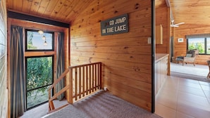 Interior de una cabaña de madera con un letrero que dice "GO JUMP IN THE LAKE", una escalera de madera y vista al exterior.
