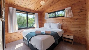 Un dormitorio con paneles de madera, una cama grande, dos ventanas con cortinas, una mesita de noche pequeña y una lámpara en el techo.