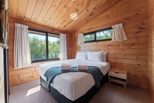 A wooden-paneled bedroom with a large bed, two windows with curtains, a small bedside table, and a ceiling light.