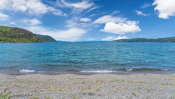 Un lago tranquilo con una playa de guijarros y colinas a lo lejos.