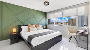 A modern bedroom with a large bed, a nightstand, a desk, and a view of the city through large windows. - BeachFront Haven/Isla Verde Prime Location/Parking (Carolina)