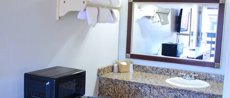 A compact hotel room with a kitchenette, a bed, a small table, and a bathroom with a sink and mirror.
