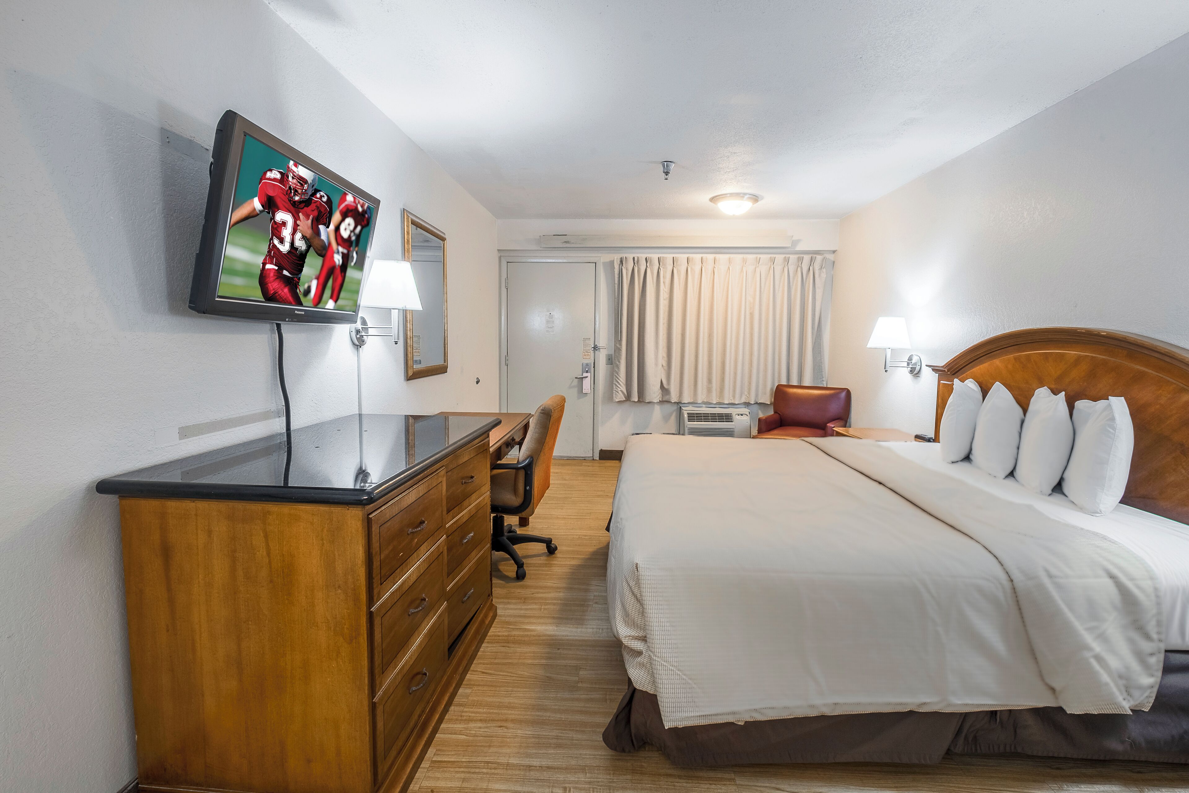 Ett hotellrum med en säng, ett skrivbord, en stol, en tv och ett fönster med gardiner.