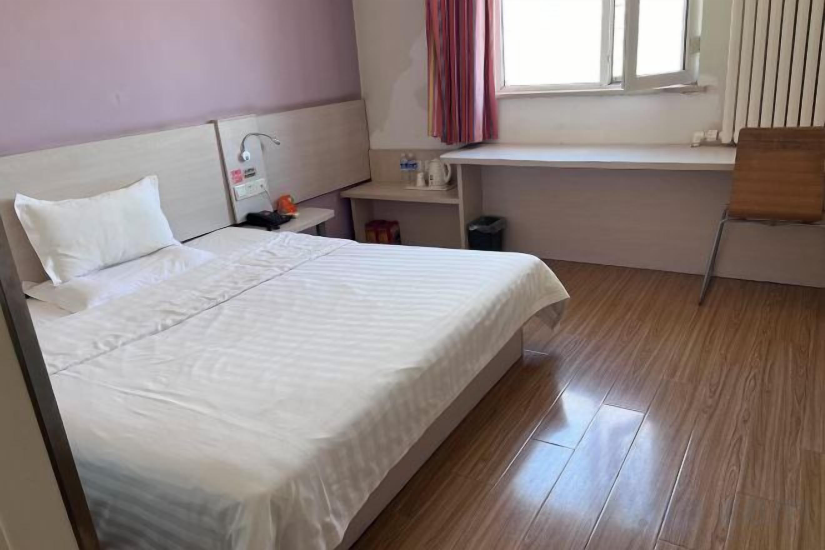A hotel room with a bed, a bedside table, a chair, a window, and a small desk with a lamp.