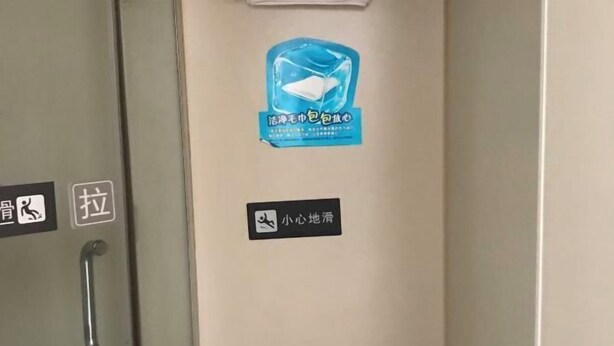 A bathroom with a handrail, a towel rack, a sign with a blue icon and text, and two rolls of toilet paper.