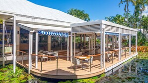 Ein überdachter Essbereich im Freien mit Tischen und Stühlen, ein Pool mit Seerosen und ein weißes Gebäude mit Wellblechdach.