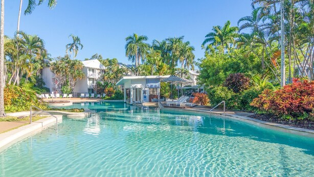Ein klares Schwimmbecken, umgeben von Palmen und Liegestühlen, mit einem Gebäude im Hintergrund.