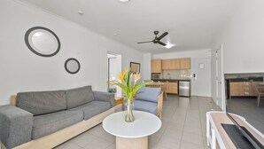 Ein modernes Wohnzimmer mit grauem Sofa, einem runden Couchtisch und einer Vase mit Blumen. Es gibt eine Küchenzeile mit Holzschränken und einer Mikrowelle.