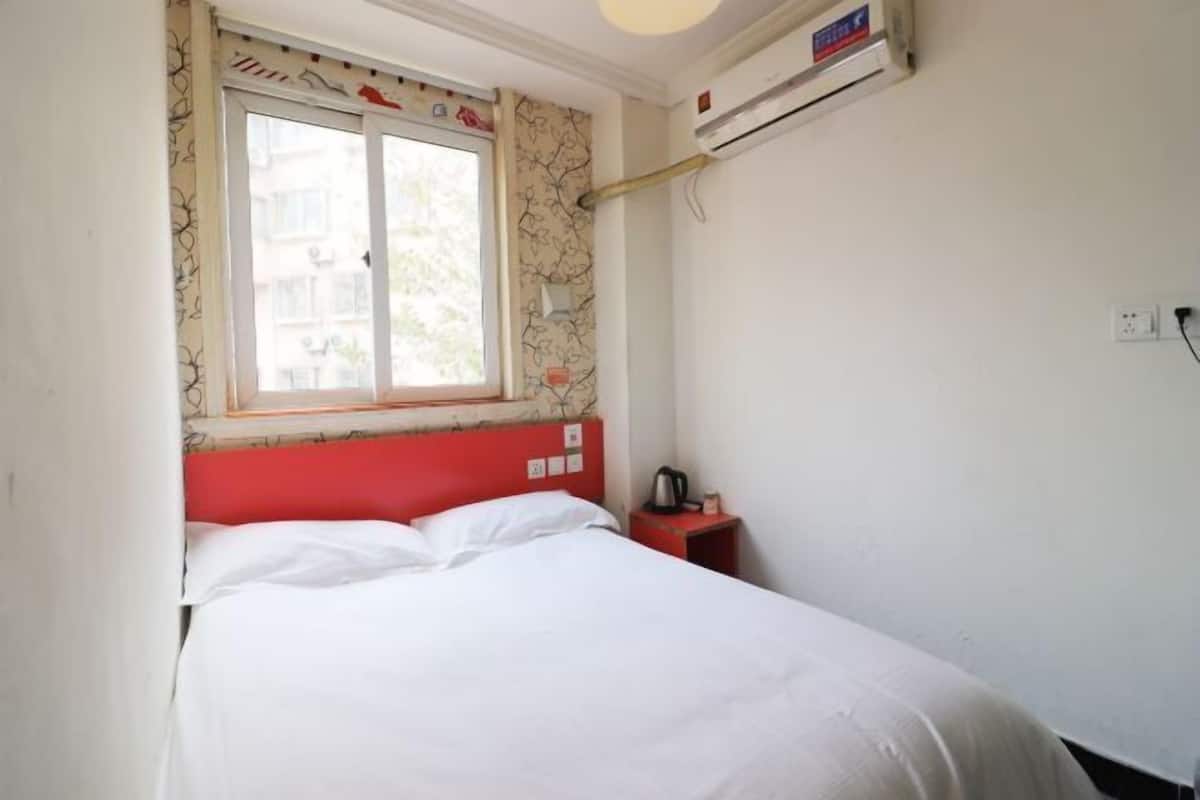 A single bed with white bedding, a red headboard, a window with patterned curtains, an air conditioning unit, and a small bedside table with a kettle.