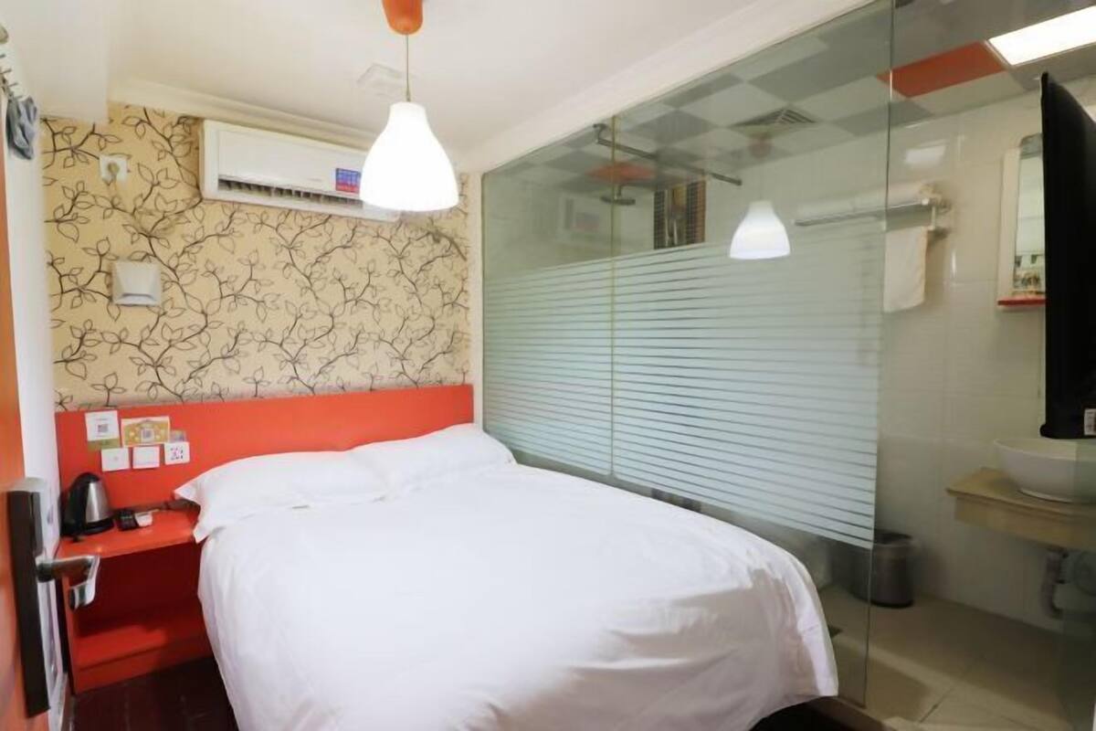 A hotel room with a bed, a red nightstand, an air conditioning unit, a glass shower enclosure, and a bathroom with a sink and mirror.