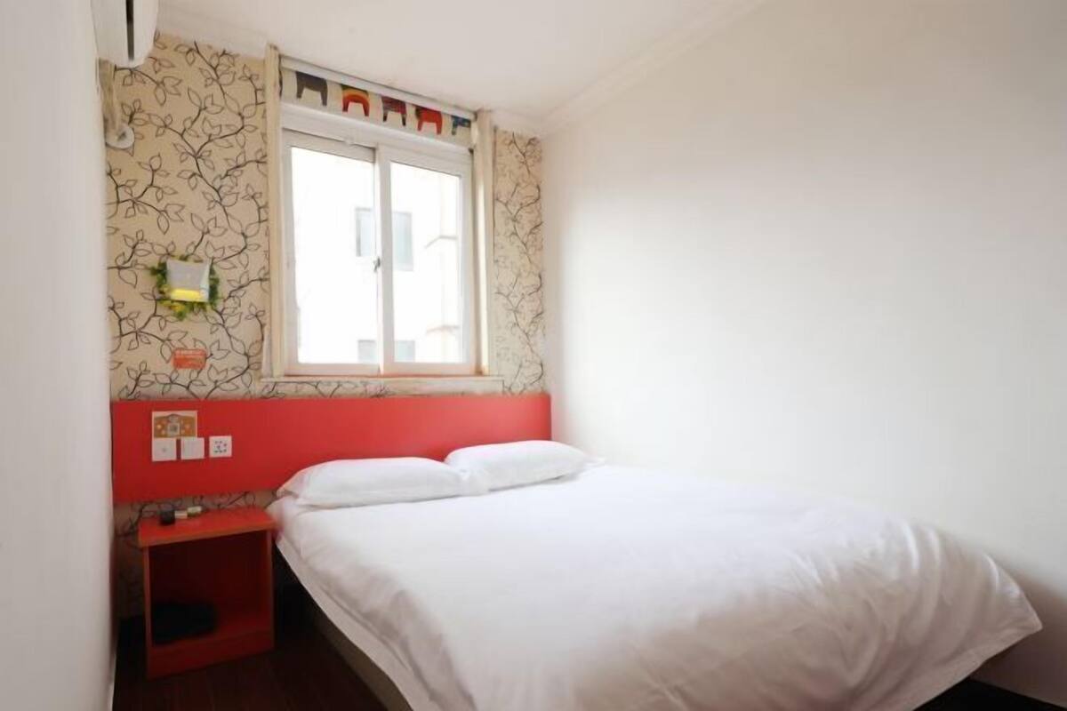 A bedroom with a bed, a window, a red headboard, and a wall with a leaf pattern.