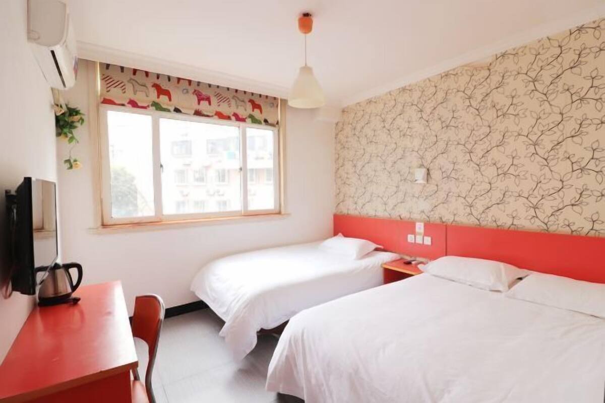 A room with two beds, a desk, a chair, a window with a curtain, and a wall with a floral pattern.
