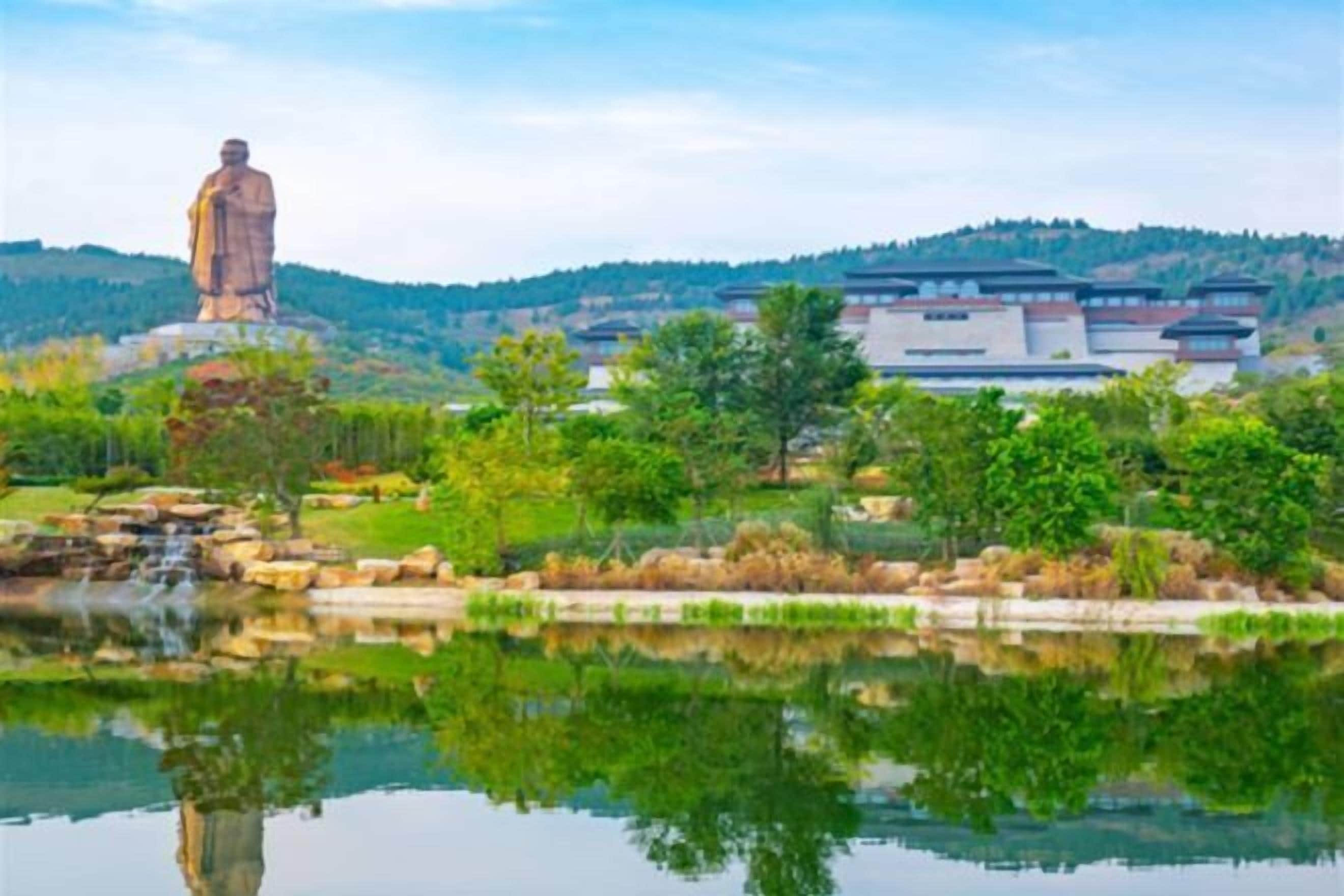 A statue on a hill with a reflection in a pond, surrounded by greenery and modern buildings.