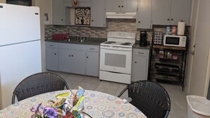 Cocina compacta con armarios blancos, estufa, microondas y una pequeña zona de comedor con sillas de mimbre.