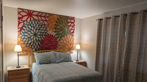 Un dormitorio con cama, dos mesitas de noche con lámparas, un tapiz decorativo en la pared y una cortina.