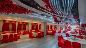 Un salón de banquetes con mesas redondas y sillas rojas, manteles blancos y un techo con follaje rojo y luces colgantes.