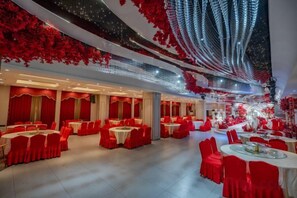 Un salón de banquetes con mesas redondas y sillas rojas, manteles blancos y un techo con follaje rojo y luces colgantes.