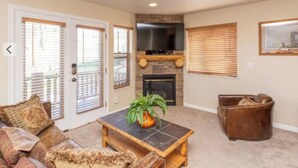 A living room with a fireplace, a television mounted above it, a sofa, a coffee table, and a brown leather chair. - Charming 3-bedroom condo in pleasant Eden with AC, fitness room, WiFi (Eden)