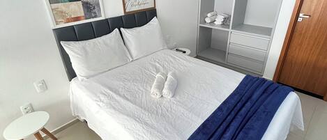Cama bem arrumada com roupa de cama branca e manta azul, mesa de cabeceira e guarda-roupa com espelho.