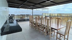 A covered outdoor dining area with a long table and chairs, a black countertop, and a view of a building in the background. - Melhores Flats - Paraíso à beira-mar (João Pessoa)