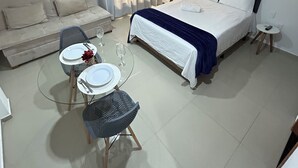 A hotel room with a bed, a sofa, a dining table with chairs, and a small round table with a remote control. - Melhores Flats - Paraíso à beira-mar (João Pessoa)