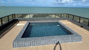 A rectangular pool with a tiled border, surrounded by a wooden deck and a glass railing. - Melhores Flats - Paraíso à beira-mar (João Pessoa)