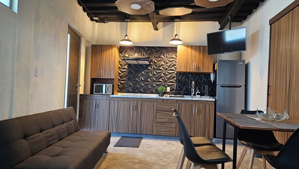 Una cocina moderna con armarios de madera, microondas integrado y refrigerador de acero inoxidable. Cuenta con un comedor con mesa y sillas, y un sofá al frente.