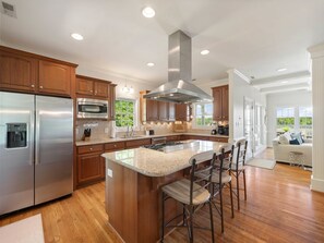 A modern kitchen with stainless steel appliances, a large island with bar stools, wooden cabinets, and a view of the outdoors through large windows. - Lake Gaston Main Lake Home, expansive view  and water toys! (Boydton)